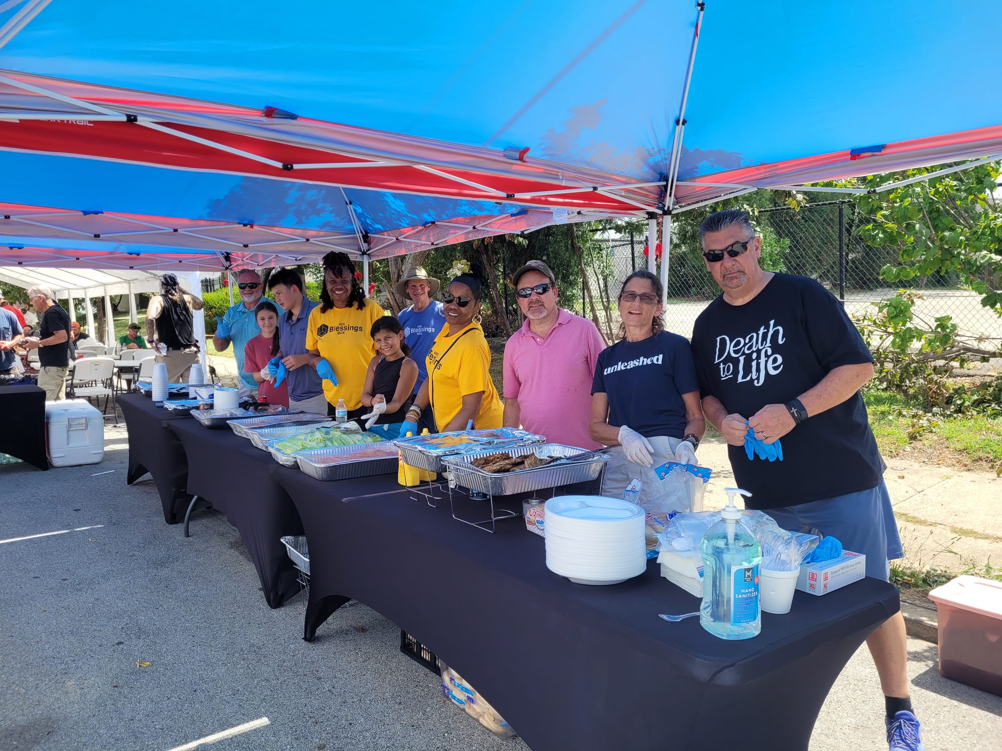 Volunteers in His Blessings Box shirts serving trays of food along tables under red and blue tents at a community day