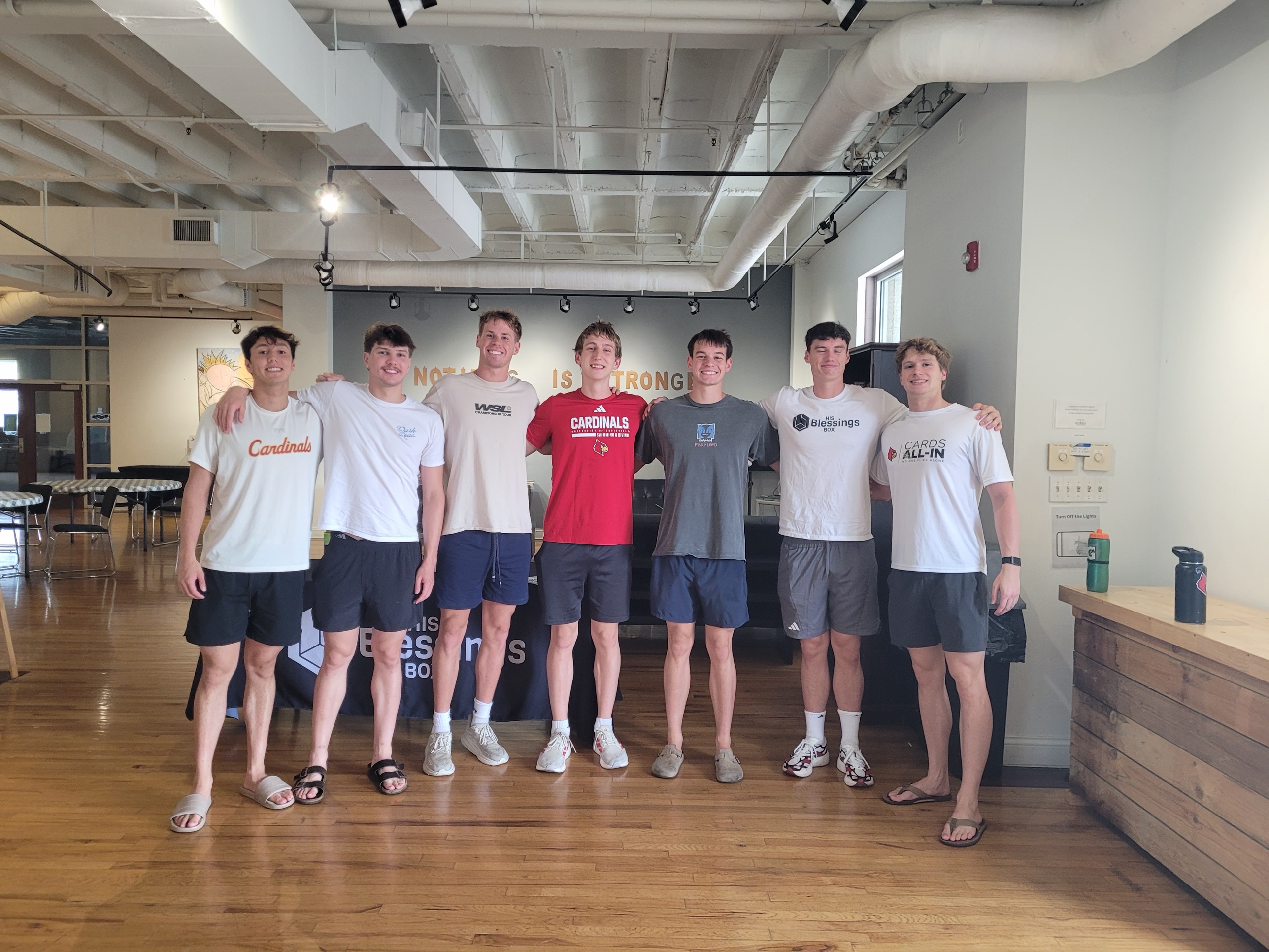 Seven smiling young men standing arm in arm in front of a His Blessings Box banner in a bright room with wood floors